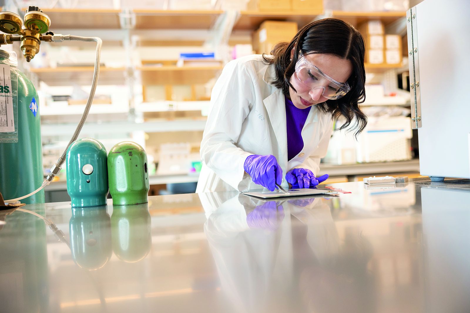 A Lehigh bioengineering researcher in a lab coat and safety glasses working at a bench beside gas cylinders