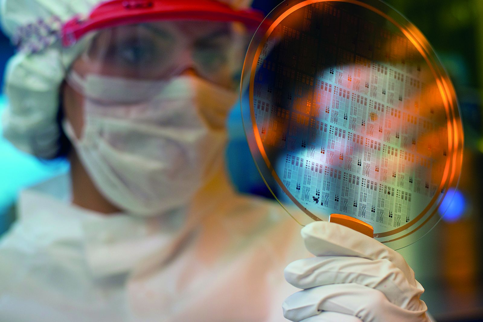 A gloved researcher in cleanroom dress holding a patterned semiconductor wafer up to the light