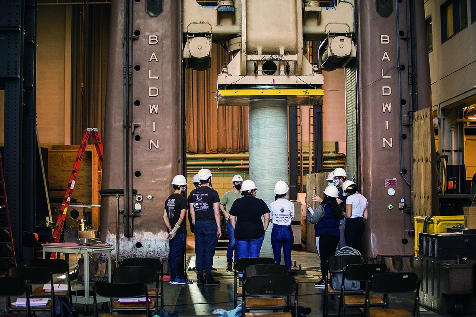 Lehigh students in hard hats gathered at the Baldwin universal testing machine in Fritz Engineering Laboratory