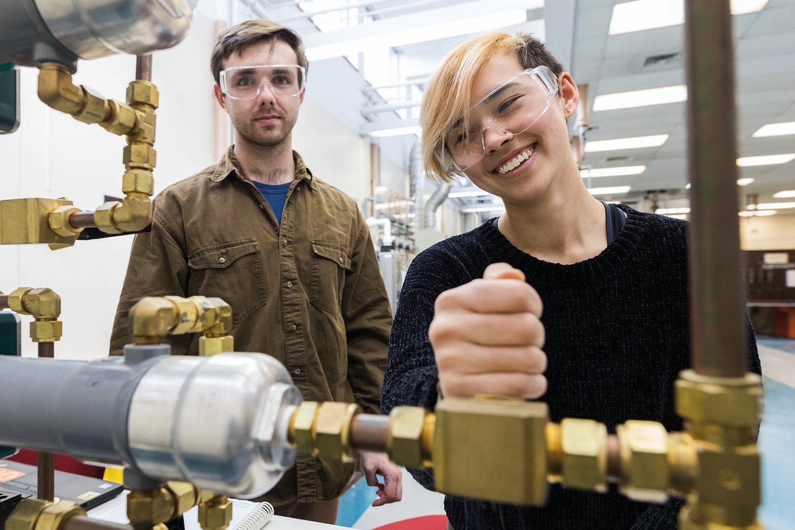 Two Lehigh engineering students in safety glasses at a laboratory rig of brass pipe fittings, one smiling as she turns a valve