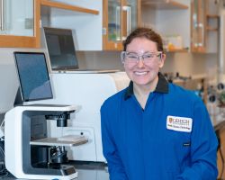 Photo of Whitney Blocher McTigue, smiling and posed in her lab and blue lab coat